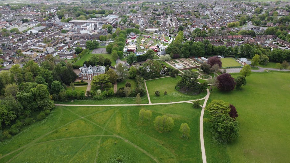 An aerial view of Clitterhouse Park in Cricklewood showing well-maintained lawns, paved walking paths, and diverse trees with lush green and purple foliage. The park features neatly trimmed grass area