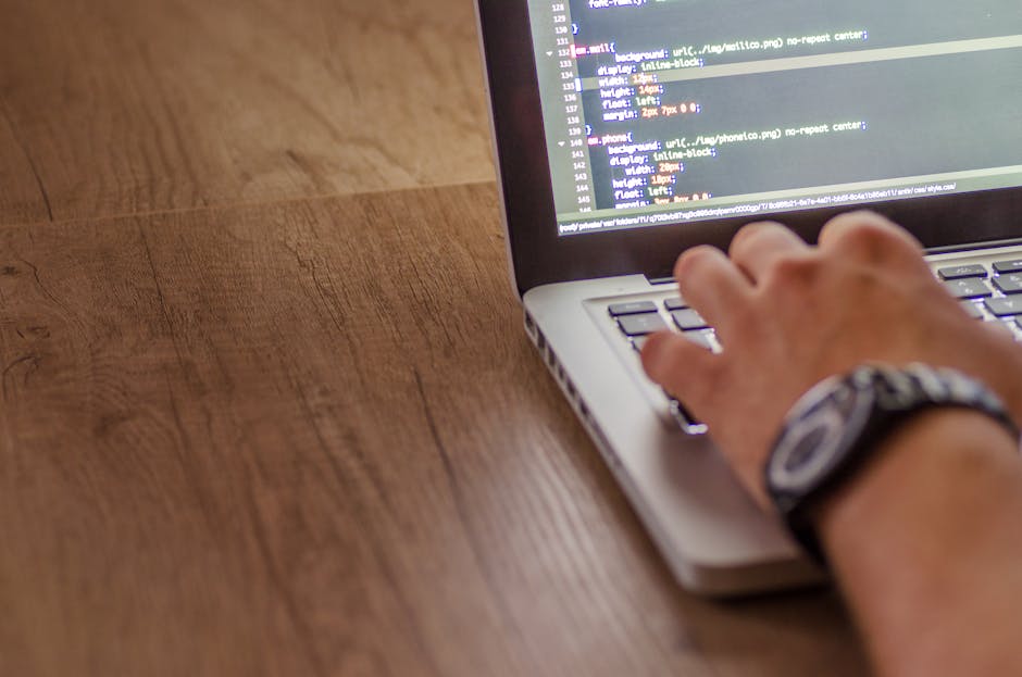 A close-up of a person's hand typing on a silver laptop placed on a wooden desk with a smooth, polished surface. The laptop's screen displays lines of code or text in a code editor with a dark backgro