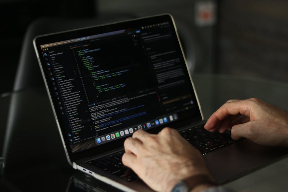 A close-up view of a person's hands operating a laptop on a dark table, displaying a code editor with colorful lines of programming code. The background is blurred, emphasizing the focus on the hands 